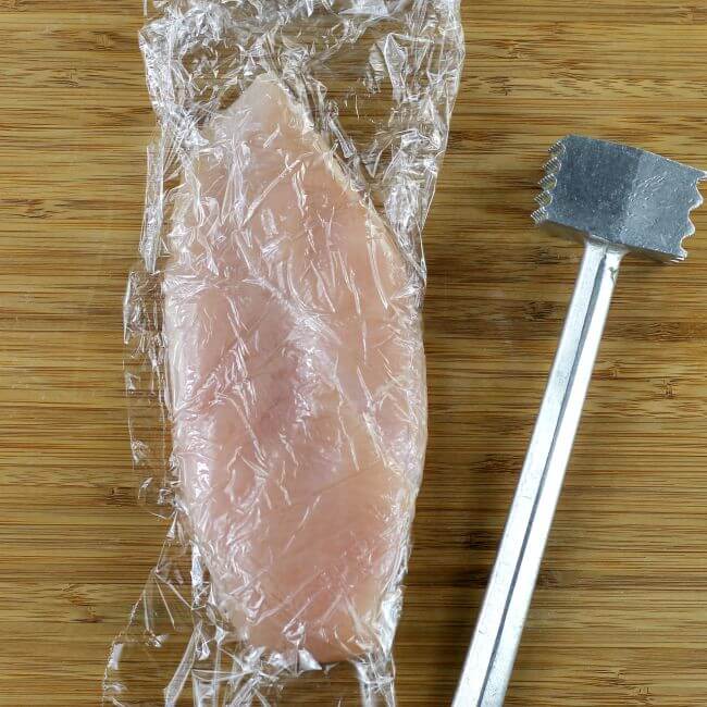 Chicken breast wrapped in plastic with mallet on wood cutting board.
