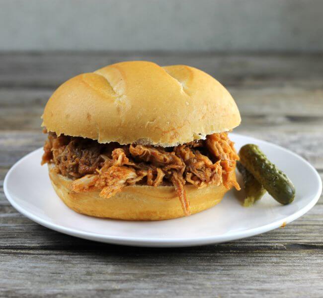 BBQ pork sandwich on a white plate with pickles on the side.