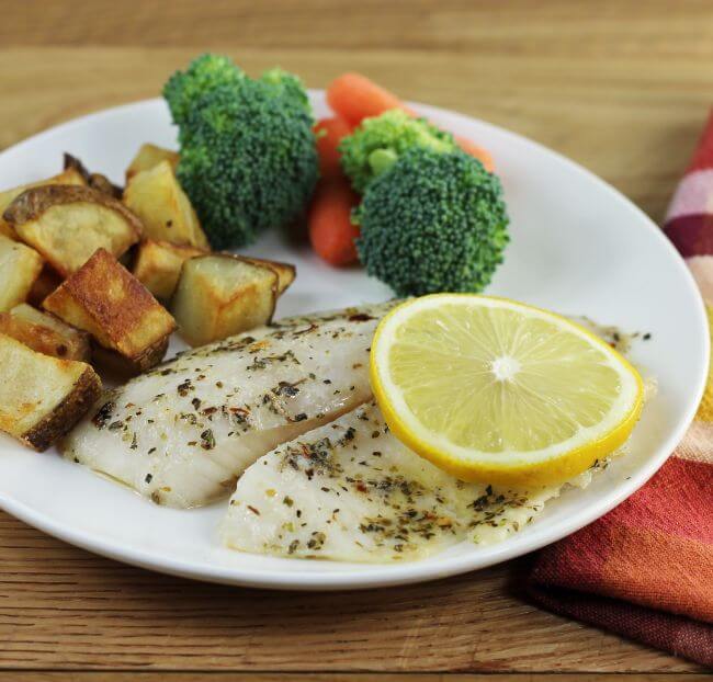 Side angle view of a tilapia fillet with lemon,  potatoes , and vegetables.