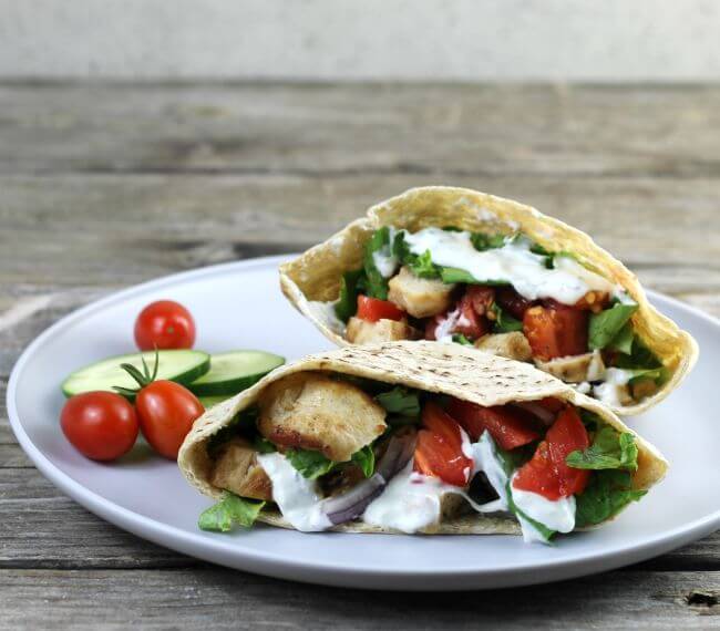 A pita stuffed with chicken, tomatoes, onion, and lettuce with tomato and cucmumber on the side