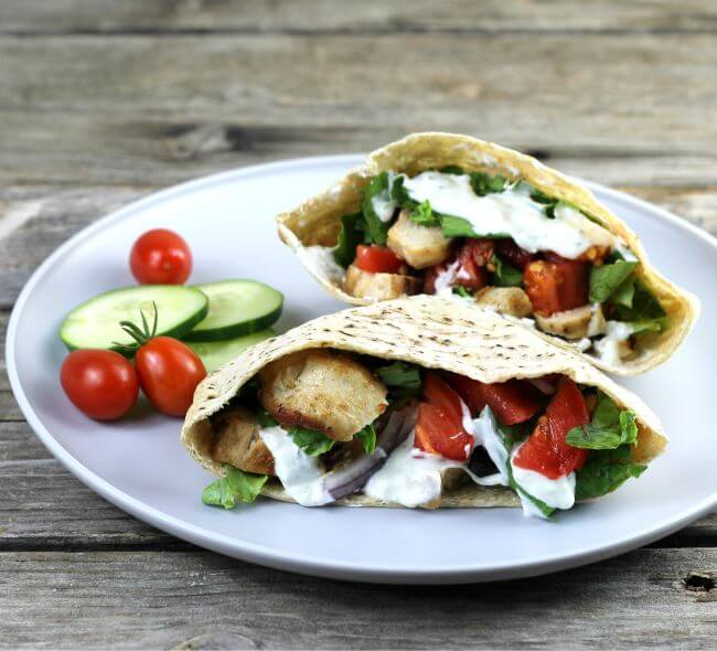 A pita stuffed with chicken, tomato, and lettuce with cherry tomatoes and cucumber on the side.