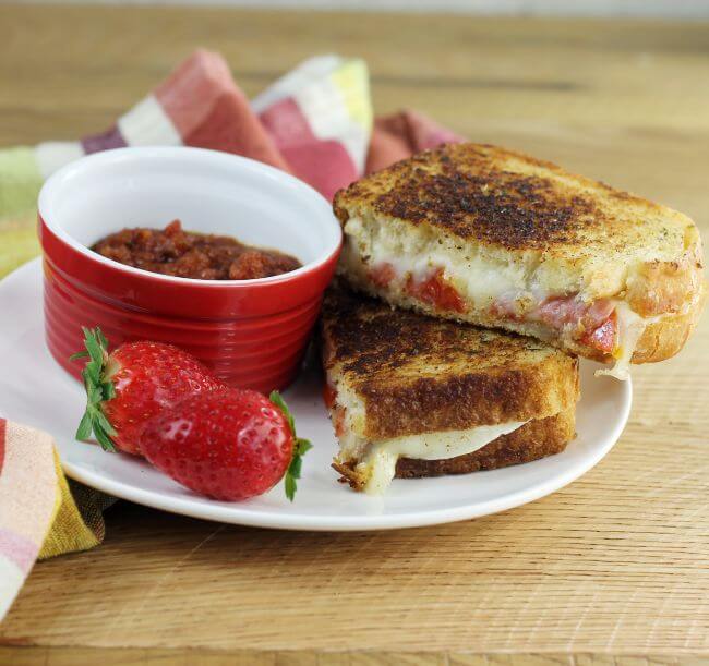 Side view of a pizza grilled cheese with a couple of strawberries and a bowl of pizza sauce.