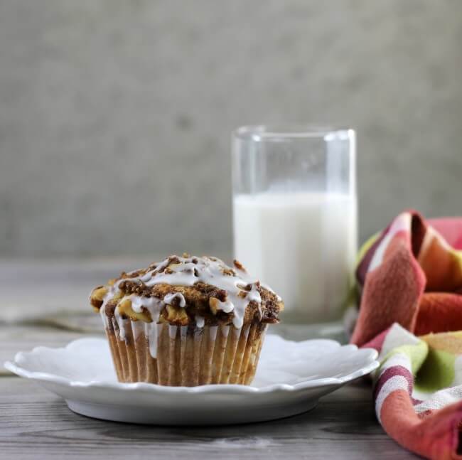 Side view of a apple steusel muffin on a white plate with a glass of milk.