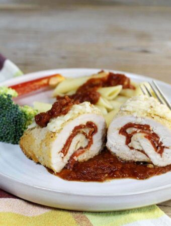 A side angle view of a chicken breast rolled up with pepperoni and cheese on a plate with veggies and pasta.