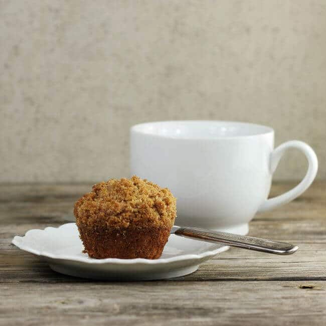 Side view of a muffin on a white plate with a coffee cup behind.