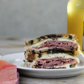 A pastrami sandwich on a white plate with a bottle on one side and a napkin on the other side.