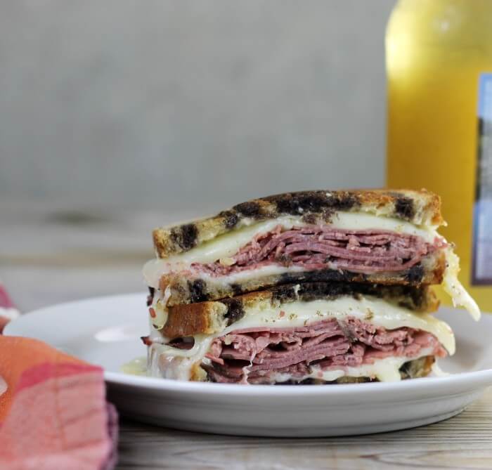 A pastrami sandwich on a white plate with a bottle on one side and a napkin on the other side. 