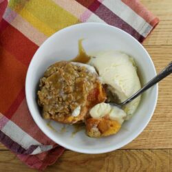 A bowl with a baked peach with crumble on the top and a scoop of ice cream with a spoon.