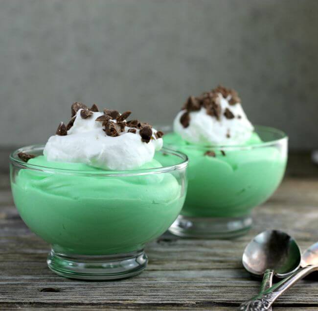 Two bowls if green pudding with whipped cream and chopped chocolate on top.