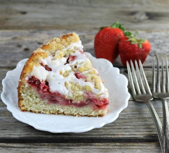 Side angle view of a slice of coffee cake with forks and strawberries on the side.