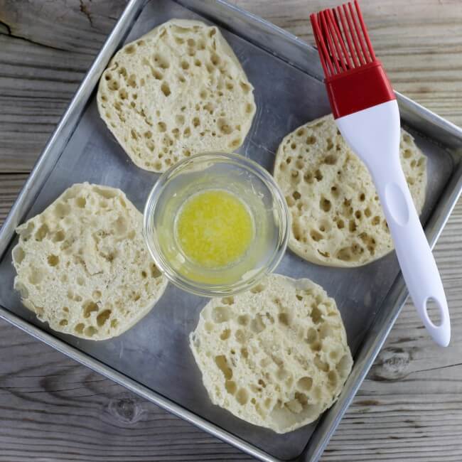 Split English muffins in a baking pan with a small bowl of melted butter with a pastry brush.