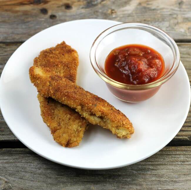 Side angle view of chicken strips with a cup of salsa on a white plate.