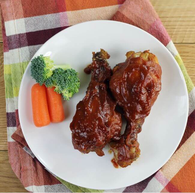 Over looking a plate with two BBQ chiccken drumsticks, with broccoli and carrot sticks.