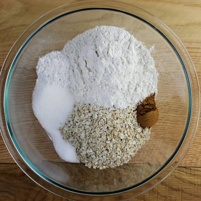 A glass bowl with flour, oats, sugar, baking powder, salt, and cinnamon in it.