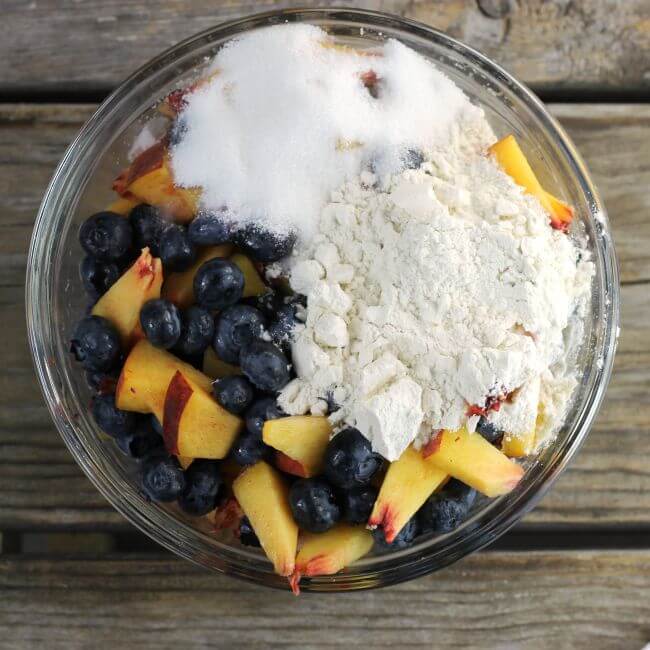 A bowl ov cut up peaches and blueberries with flour and sugar on top.