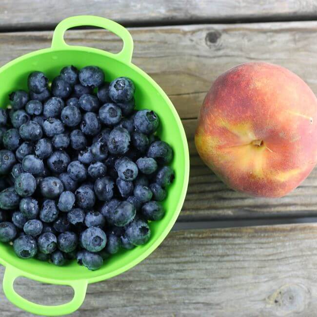 Blueberries in a green colander with a peach on the side.