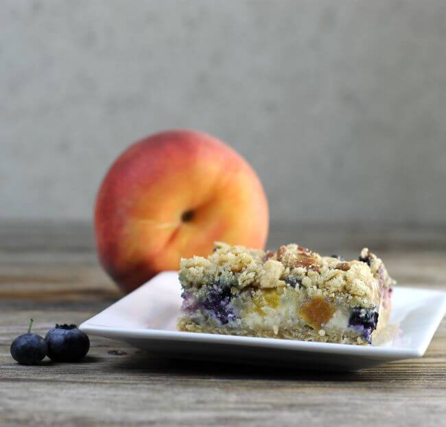 A side view of a blueberry peach bar with a peach in the back ground and blueberry.