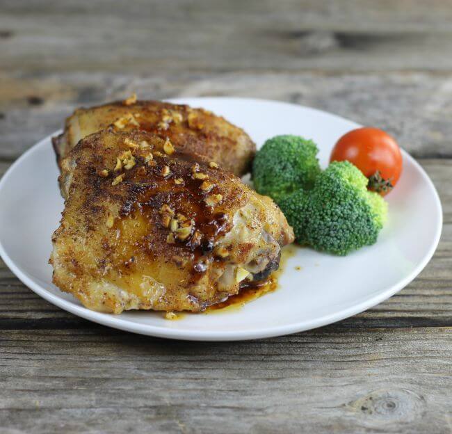 Two chicken thighs on a white plate with broccoli and a cherry tomato.