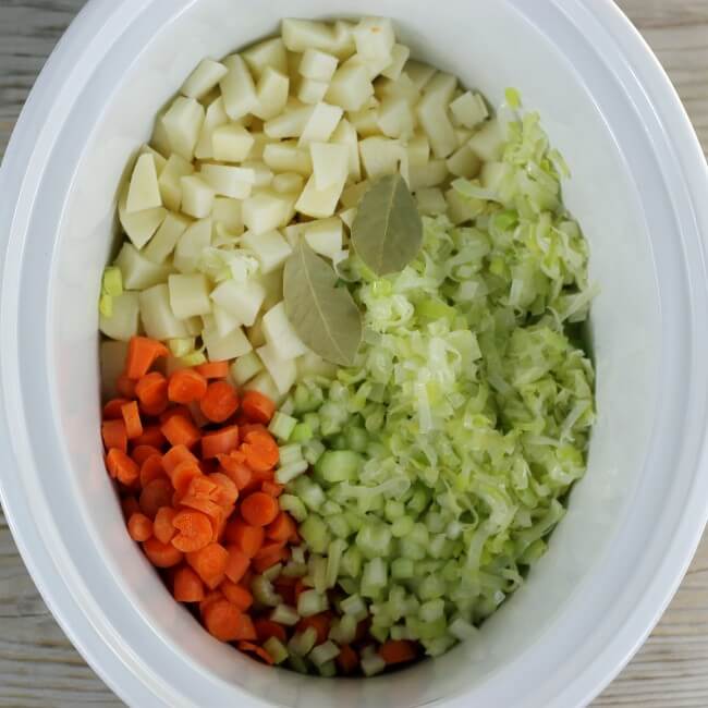 Celery, carrots, potatoes, leeks, and bay leaves are added to the slow cooker.