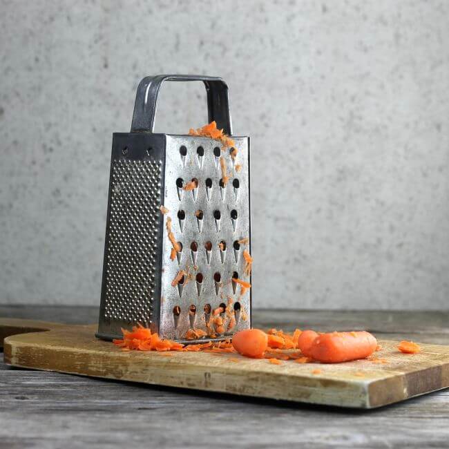 Box grater with carrots on a wooden bread board.