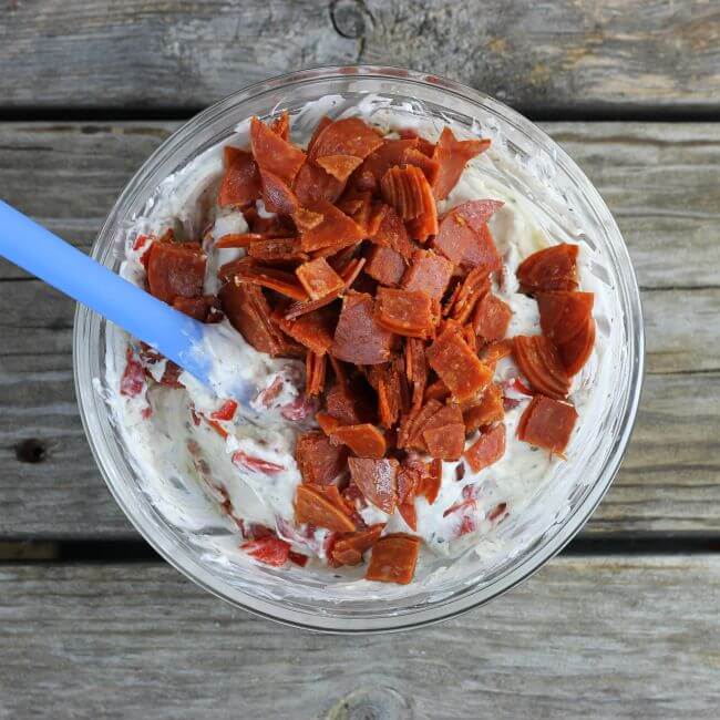 Glass bowl with spatula in the bowl with cream cheese dip, pepperoni and roasted red peppers.