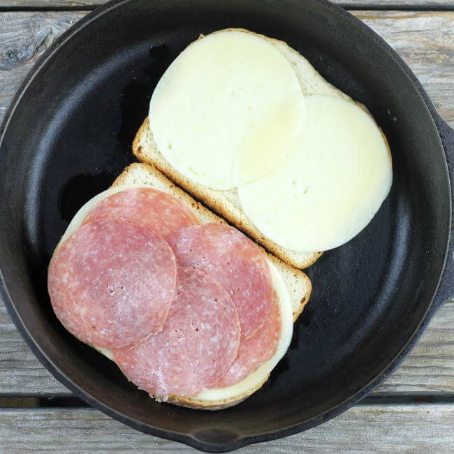 2 slices of bread with cheese and salami in skillet.