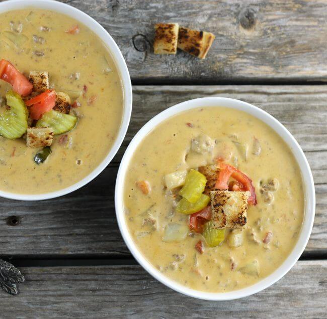 Two bowls of cheeseburger soup. with tomatoes, pickles, and croutons on top.