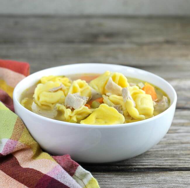 Chicken tortellini soup in a white bowl with a plaid napkin on the side.