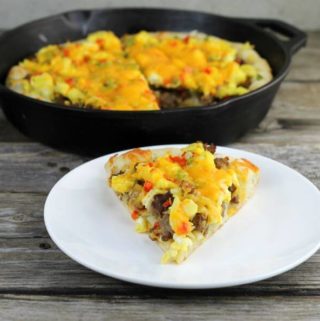 Sausage breakfast pizza on a white plate with the cast-iron skill with the remaining pizza behind.