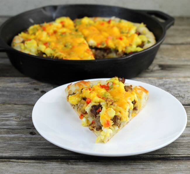 Sausage breakfast pizza on a white plate with the cast-iron skill with the remaining pizza behind.