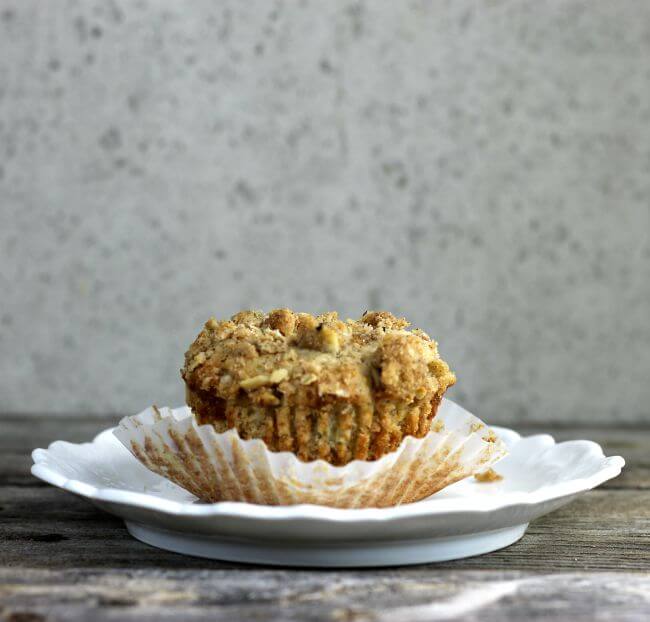 Beat banana muffin with the paper pulled down on the muffin which is sitting on a white plate.