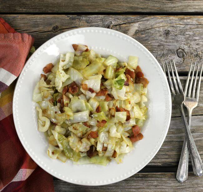 Overhead view of fried cabbage with bacon in a white bowl with forks and napkin on the sides.