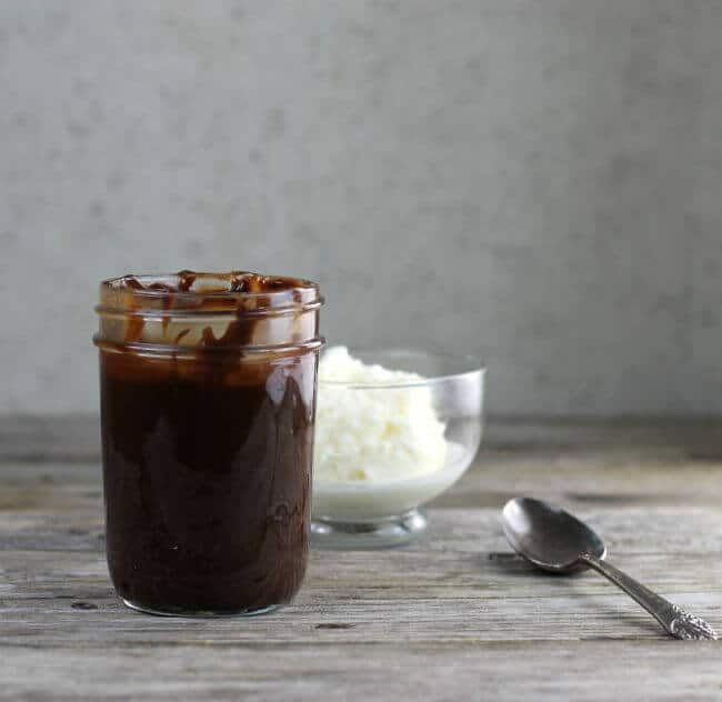 A jar of hot fudge with a bowl of ice cream behind it.