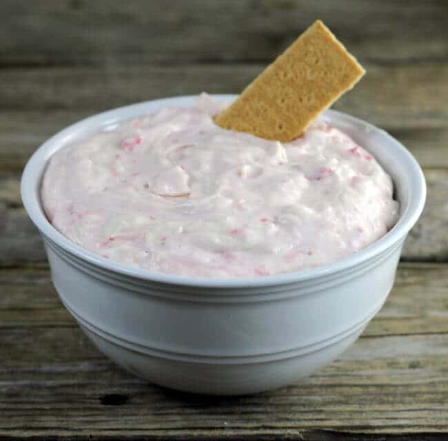Awhite bowl of cheesecake dip with a graham cracker sticking out of the dip.