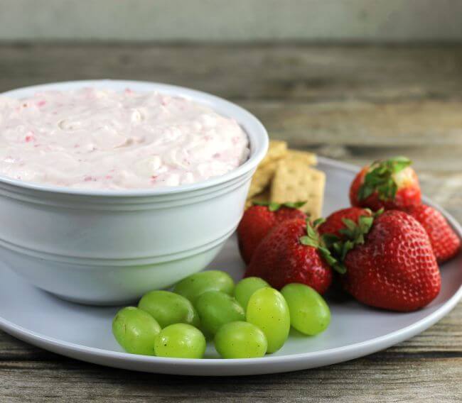 Side view of a bowl of cheesecake dip on a plate with grapes and strawberries.