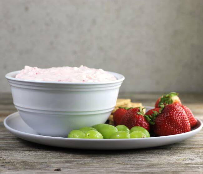 Side view of a bowl of cheesecake dip on a plate with fresh fruit.