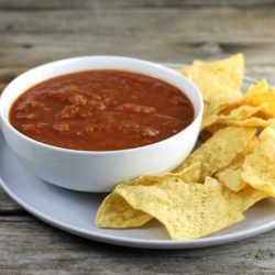 Side view of a bowl of salsa in a white bowl with chip around the bowl on a gray plate.