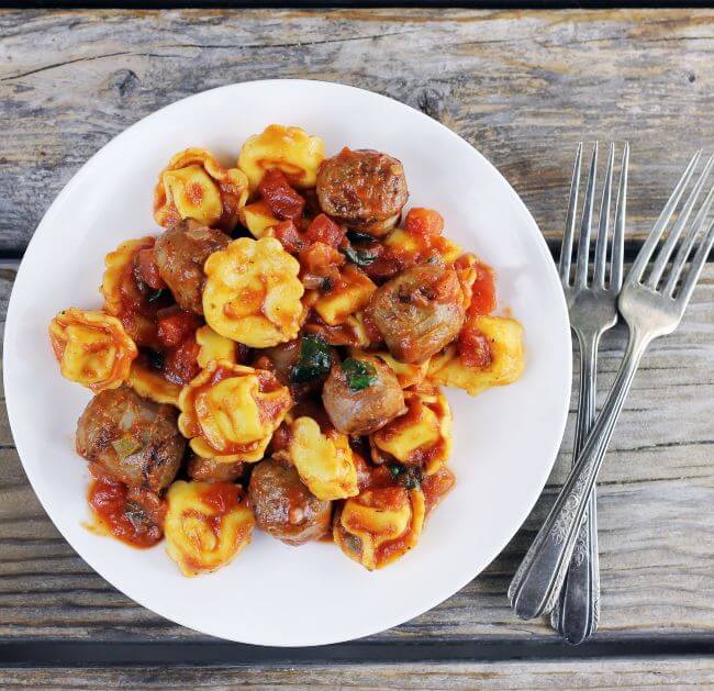 Looking down at a plate of Italian sausage and tortellini with forks on the side.