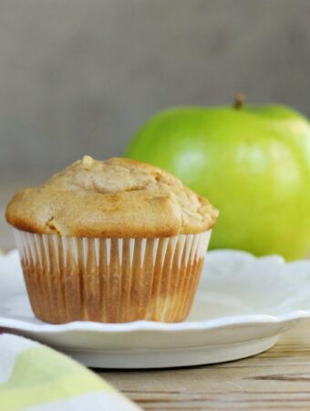 A side view of a white plate with an apple muffin with an apple in the back.